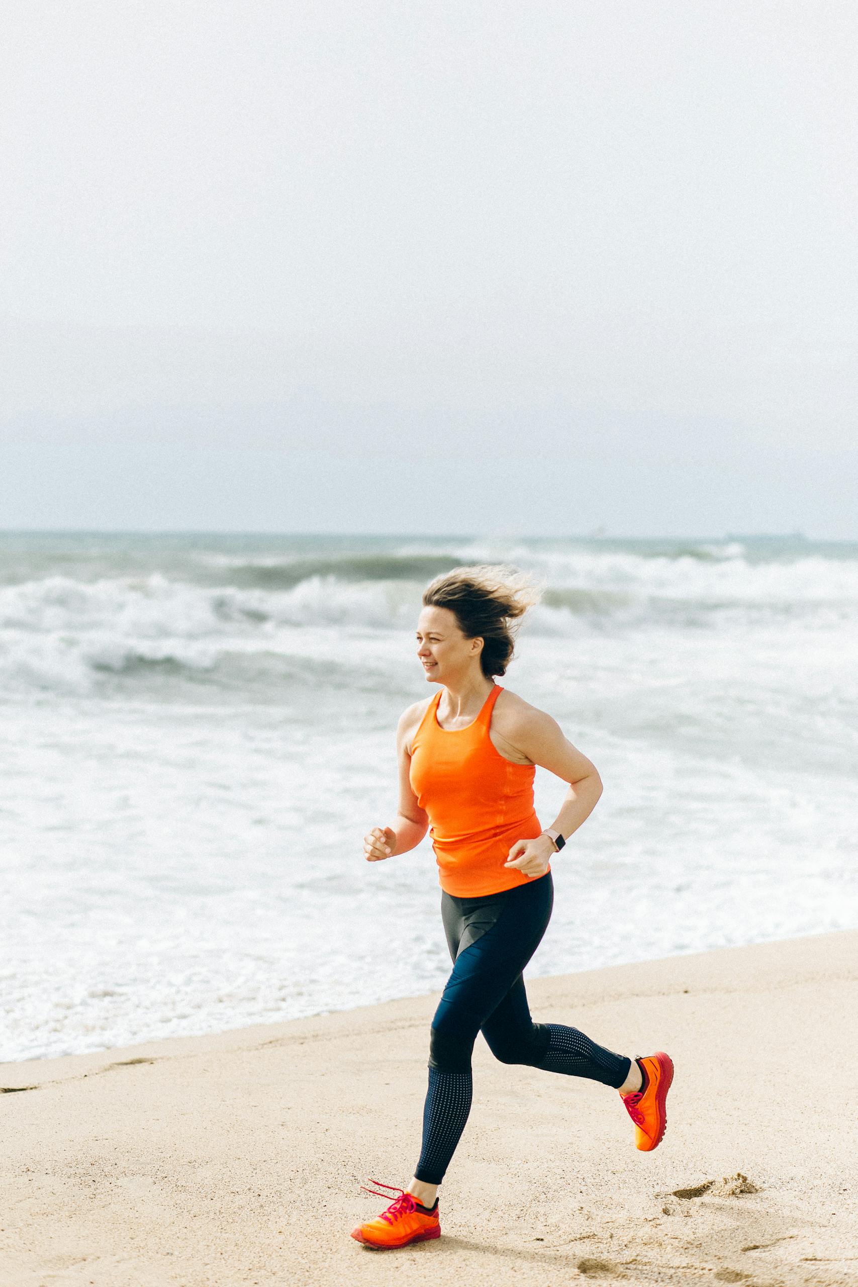 Caucasian woman jogging on the beach in orange tank top, embodying active lifestyle and fitness.